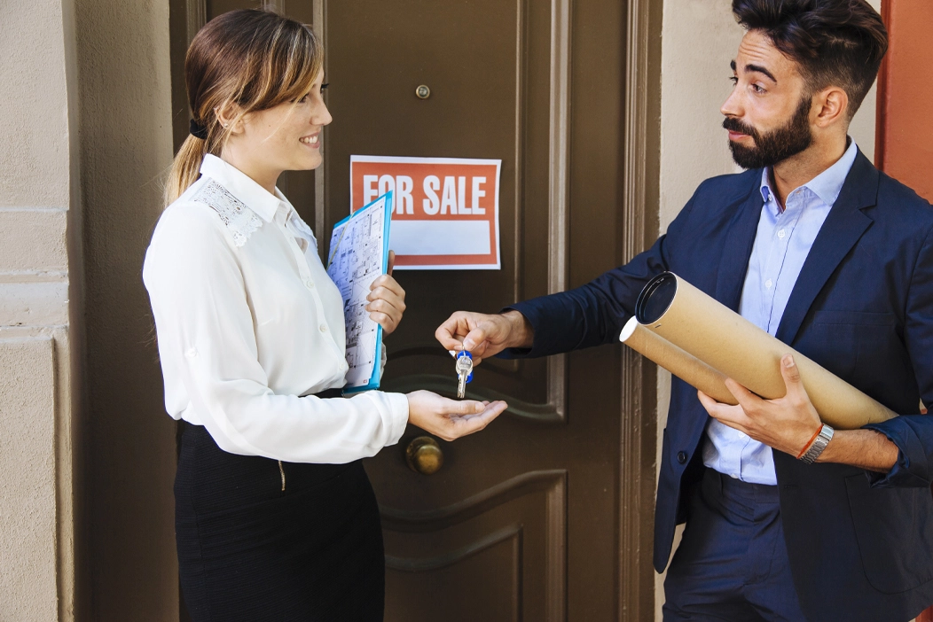 Guy giving key to lady and there is a sign on the door showing For Sale
